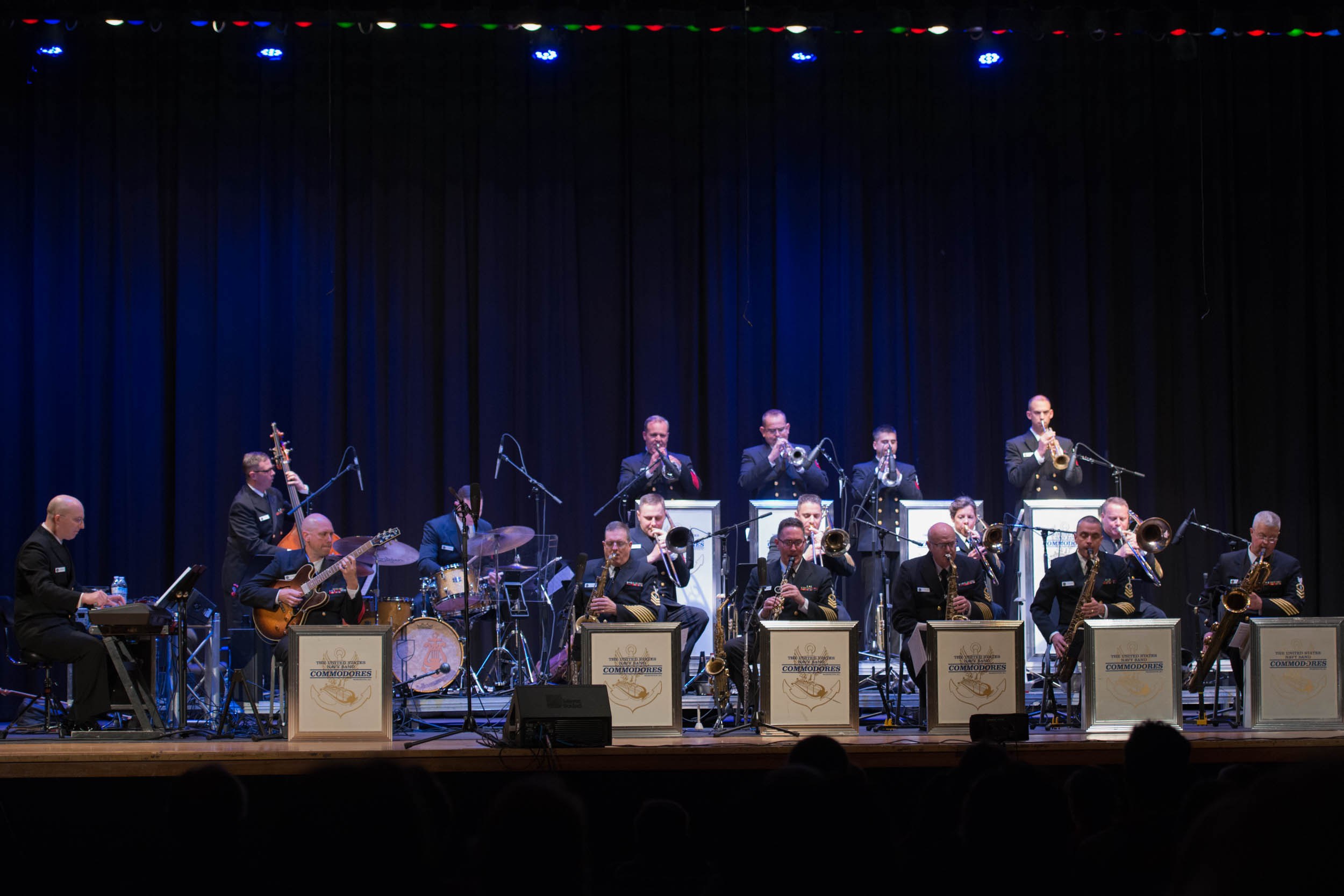 Eine Gruppe von Musikern tritt auf einer hell erleuchteten Bühne auf, die als "The New Orleans Jazz Band" beschriftet ist, mit Mikrofonen und Ständern, während Zuschauer von unten zuschauen.