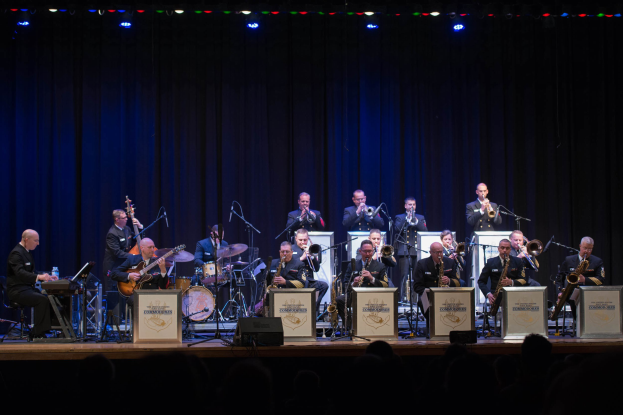 Eine Gruppe von Musikern tritt auf einer hell erleuchteten Bühne auf, die als "The New Orleans Jazz Band" beschriftet ist, mit Mikrofonen und Ständern, während Zuschauer von unten zuschauen.