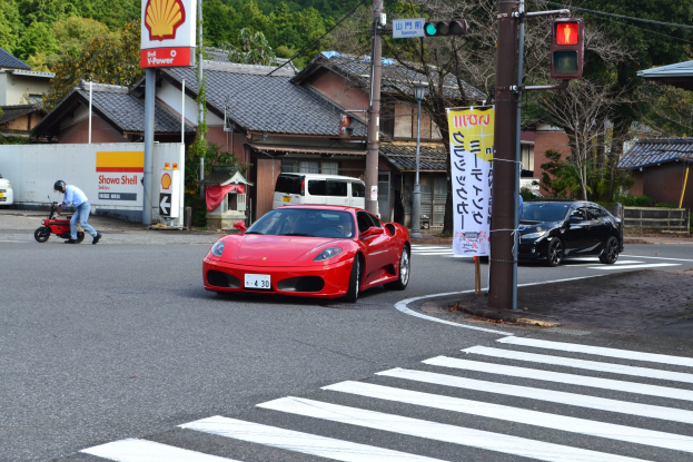 Roter Sportwagen fährt auf einer Straße an einer Ampel vorbei, mit einer Person auf einem Roller im Vordergrund und Gebäuden, Bäumen und anderen Fahrzeugen im Hintergrund.