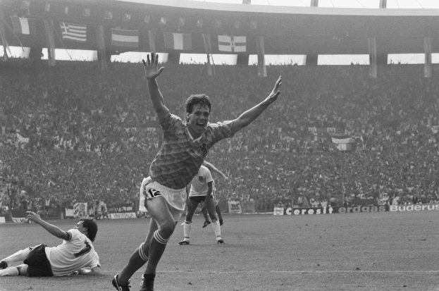 Schwarzes und weißes Foto eines Fußballspielers, der ein Tor feiert, umgeben von Mannschaftskameraden und Betreuern auf dem Feld, mit Stadiontafeln, Fahnen und Pfosten im Hintergrund unter einem klaren Himmel.