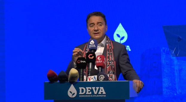 Ein Mann in einem Mantel und einem Schal steht an einem Podium mit Mikrofonen und hält eine Rede vor einem Publikum mit einer Tafel, auf der der Text "deva ceo sagt, dass es keine gute Idee für die Zukunft ist" zu sehen ist.