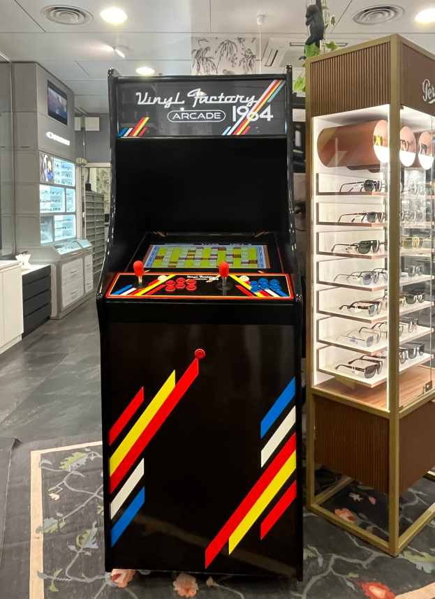 Ein Geschäft mit einem zentralen Retro-Arcade-Automaten, einem Gestell mit Gläsern auf der rechten Seite und Hintergrundelementen wie Schränken, einem Fernseher und anderen Gegenständen, wobei Deckenleuchten für eine Retro-Atmosphäre sorgen.