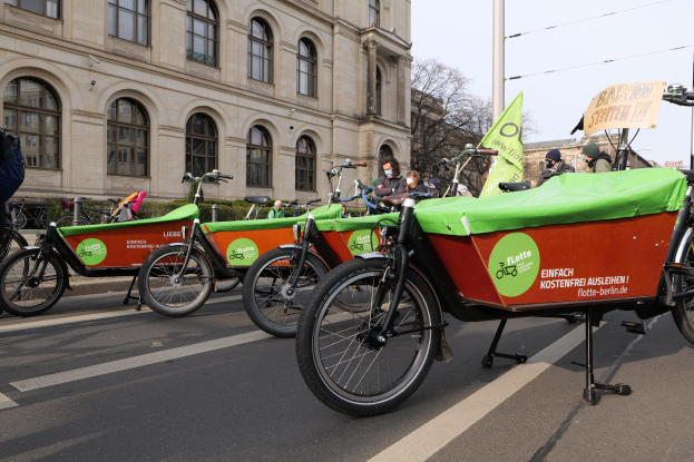 Eine Gruppe von Fahrrädern, die auf einer Straße mit einer Person in der Nähe geparkt sind, Gebäude und Bäume im Hintergrund und ein Banner, das eine Fahrrad-Sharing-Kampagne anzeigt.