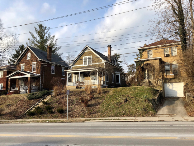 Reihe von Häusern mit Fenstern, Türen, Säulen, Geländern, Stufen, Pflanzen und einem Pfahl mit einer Tafel entlang einer Straße, mit Bäumen und Gras davor, unter einem bewölkten Himmel.