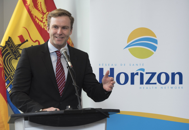 Ein Mann mit einem freundlichen Gesichtsausdruck in einem schwarzen Blazer, einem weißen Hemd und einer roten Krawatte steht an einem Rednerpult mit einem Mikrofon, während er als CEO spricht, mit einem Banner im Hintergrund.