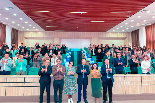 Eine Gruppe von Menschen steht vor einer Menge, einige klatschen, in einem Raum mit Stühlen, Tischen, Luftballons, Vorhängen und Deckenleuchten, feiernd die Eröffnung einer neuen Schule.