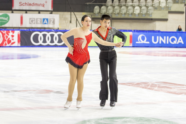 Zwei Eiskunstläufer, ein Mann und eine Frau, gleiten Hand in Hand über eine Eisbahn mit Zuschauern und Absperrungen im Hintergrund.