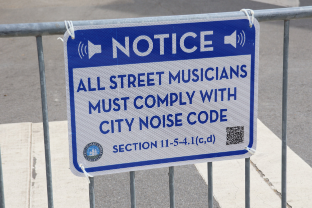 Anschlagtafel an einem Zaun mit einem Schild, auf dem "Anschlag: Alle Straßenmusiker müssen die Stadtlärmvorschriften einhalten" steht, und eine Straße im Hintergrund.