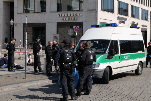Eine Gruppe von Polizisten steht vor einem Starbucks-Kaffeehaus, rechts ein Van und links einige Menschen; im Hintergrund sind ein Gebäude, ein Schild, ein Laternenpfahl und ein Zaun zu sehen.