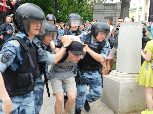 Eine Gruppe von Einsatzpolizisten in Ausrüstung, die vor einer Menge weglaufen, einige mit Helmen und Waffen, mit einer Säule im Vordergrund, Bäumen, Gebäuden und Laternen im Hintergrund und Kameras in der Menge sichtbar.