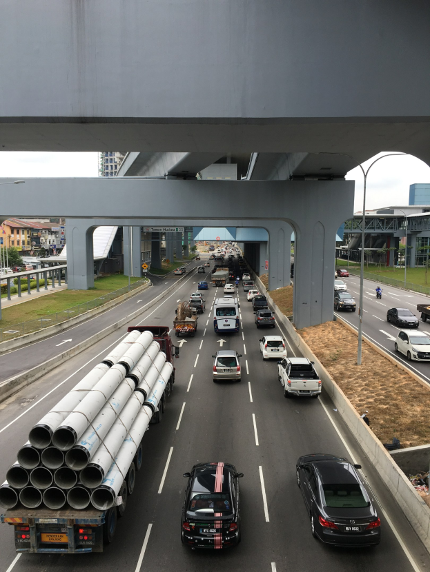 Eine vielbefahrene Autobahn mit mehreren Fahrzeugen, einer Brücke darüber, Straßenlaternen, Gras, Gebäuden, Bäumen und dem Himmel im Hintergrund.
