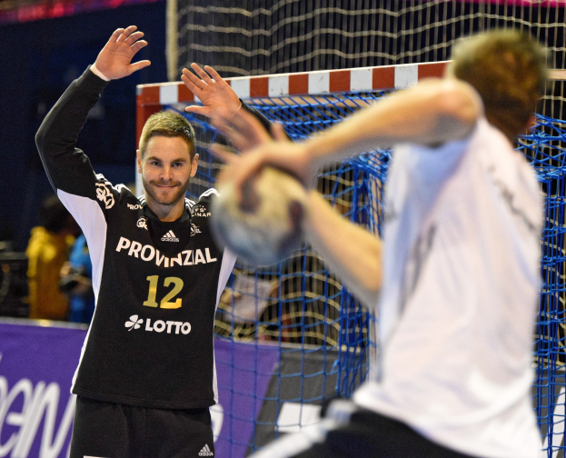 Zwei Männer spielen Handball vor einem Tor mit Netz und einem Banner im Hintergrund, in einer Stadionatmosphäre.