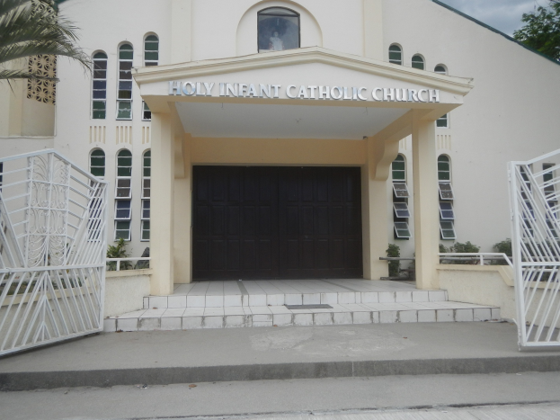 Die Heilige Kindermissionskirche, ein Gebäude mit Fenstern und einer Tür, Treppen führend zur Einfahrt, Geländern auf beiden Seiten, umgeben von Pflanzen und Bäumen, mit einem bewölkten Himmel im Hintergrund.