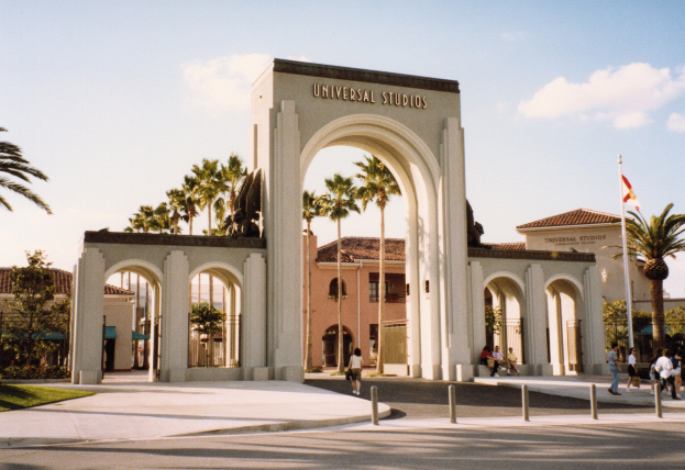 Eingang zu Universal Studios Hollywood mit einem Bogen mit Text, einer Statue, Menschen, umgeben von Grün, Gebäuden, einem Fahnenmast mit einer Flagge und einem bewölkten Himmel.