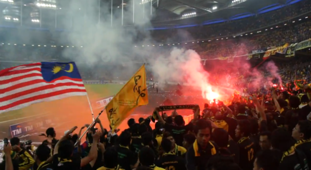 Eine große Menschenmenge in einem Stadion mit Bengalos, Fahnen und einer Kamera, mit Rauch und Schildern, auf denen 'Malaysia vs Malaysia Live Streaming' steht.