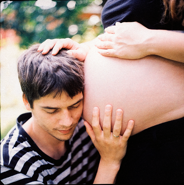 Ein Mann und eine Frau stehen nebeneinander, wobei der Mann zärtlich den Bauch der Frau berührt und auf das ungeborene Kind schaut.