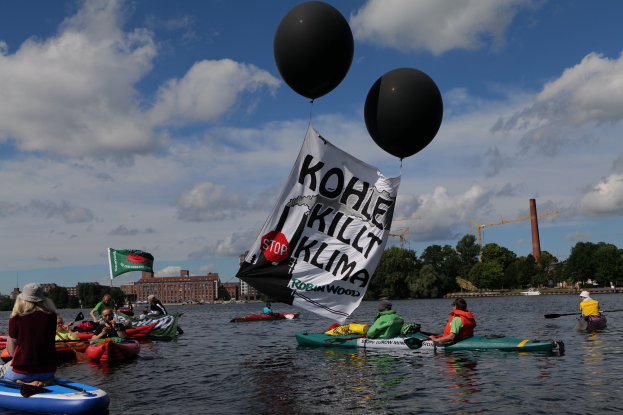 Gruppe von Menschen in Kajaks auf dem Wasser paddeln mit einem "Kohle Kill Klima"-Schild an einem Boot, haltend Paddel, vor einem Hintergrund von Bäumen, Gebäuden, Kränen und einem klaren blauen Himmel.