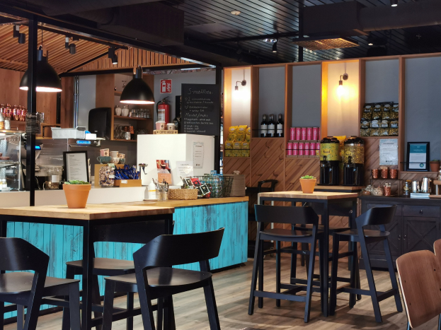 Innenansicht eines Restaurants mit Holztischen, Stühlen, einer Topfpflanze auf einem Tisch, einer Schiefertafel, einem Kühlschrank, einem Feuerlöscher, Regalen mit Flaschen, Schränken mit Gegenständen, einem Rahmenfoto an der Wand, Deckenleuchten und Lampen.