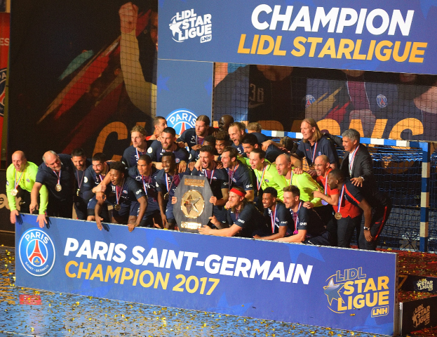 Paris Saint-Germain-Spieler feiern mit der Ligue-1-Trophäe, während sie Medaillen tragen, vor einer Tafel mit Text und Logos stehen, mit einem Netz auf der rechten Seite und einem Bildschirm im Hintergrund.