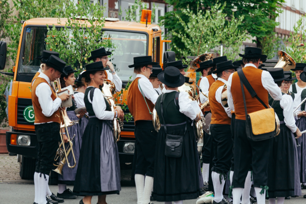 Gruppe von Menschen in traditioneller bayrischer Tracht, die eine Straße entlangmarschieren, Instrumente spielen und Taschen tragen.