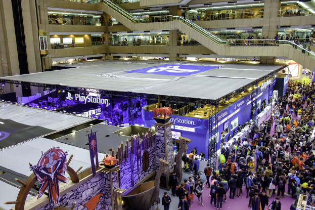 Eine große Menschenmenge in einem großen Kongresszentrum mit Rolltreppen, Geländern, beleuchteten Schildern, Bannern und anderen Anzeigen, mit einem Wasserzeichen für die weltweit größte Gaming-Convention.