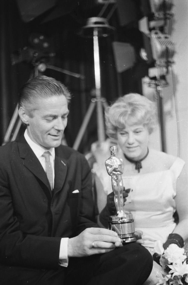 Ein Schwarz-Weiß-Bild eines formell gekleideten Mannes, der eine Oscar-Statue hält und eine Frau, die einen Blumenstrauß hält, nebeneinander sitzen.