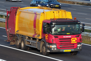 Ein gelber und roter Müllwagen auf einer Autobahn umgeben von Autos, mit einer Begrenzung und Gras an den Seiten der Straße.