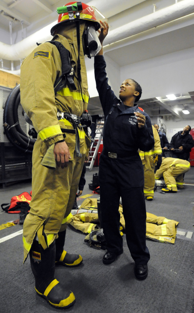 Zwei Feuerwehrleute in Uniform stehen nebeneinander, einer in einem schwarzen Kleid und Helm, der andere in gelb-schwarzer Ausrüstung, mit anderen und Equipment im Hintergrund.
