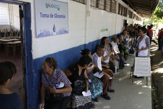 Menschen sitzen auf Bänken vor einem Gebäude, warten in der Schlange, um an einer Wahllokal in Guadalajara, Mexiko, zu wählen, mit einer Tafel an der Wand, einer Tür links und Bäumen im Hintergrund.