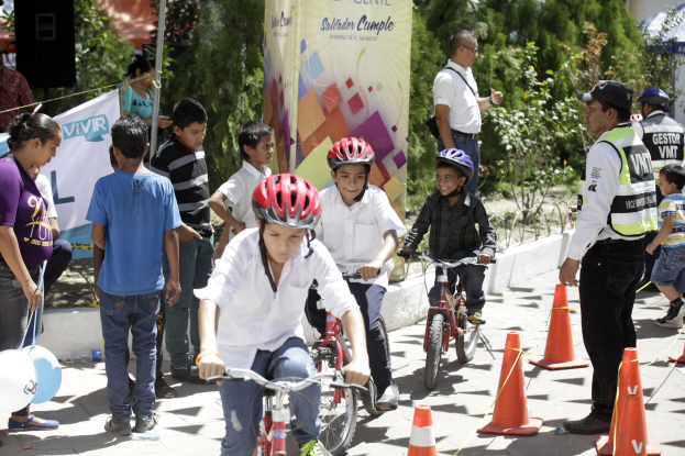 Gruppe von Kindern, die Fahrräder auf einer Straße mit Verkehrsleitkegeln fahren, einige tragen Helme, andere stehen daneben, mit einem Banner, Bäumen und Gebäuden im Hintergrund.