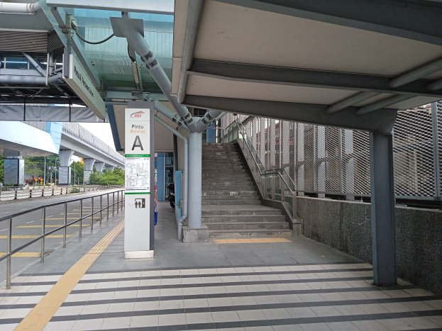 Ein Bahnsteig mit Treppe, eine Straße mit Geländer links daneben und ein Hintergrund mit Bäumen, Pfählen, Gebäuden und einer Brücke, mit Rohren und einer Tafel oben auf dem Bahnsteig.