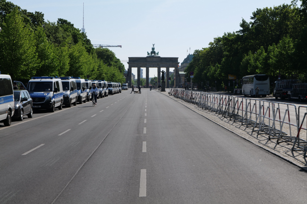 Lange Reihe von Polizeiwagen am Straßenrand vor dem Brandenburger Tor in Berlin, Deutschland, mit Menschen auf Fahrrädern und in der Nähe stehenden, Absperrungen, Bäumen und einem Bogen mit Statuen im Hintergrund.