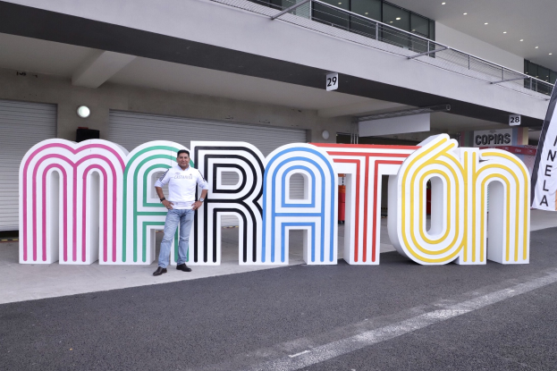 Ein Mann steht am Start eines Marathons vor einem großen "Marathon"-Schild, mit einer Fahne rechts daneben und einem beleuchteten Gebäude im Hintergrund.