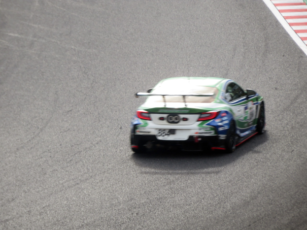 BMW M4 GT3 Rennwagen auf einer Rennstrecke mit einem Rad im Vordergrund und einer Wand mit Text auf der rechten Seite.