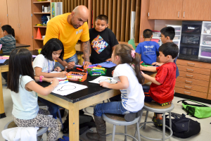 Eine Gruppe von Kindern sitzt um einen Tisch in einem Klassenzimmer, mit einem Mann davor, umgeben von Papieren, Stiften und anderen Gegenständen, mit Taschen auf dem Boden und Schränken und Regalen im Hintergrund.