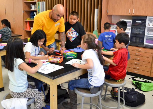 Eine Gruppe von Kindern sitzt um einen Tisch in einem Klassenzimmer, mit einem Mann davor, umgeben von Papieren, Stiften und anderen Gegenständen, mit Taschen auf dem Boden und Schränken und Regalen im Hintergrund.