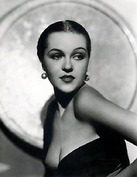 Ein Schwarz-Weiß-Foto einer Frau in einem langen Kleid im Stil der 1940er Jahre mit hohem Kragen und langen Ärmeln, gestylten Haaren, dezentem Make-up und einem selbstbewussten Ausdruck, die direkt in die Kamera blickt, mit einem unscharfen Hintergrund und Text unten.