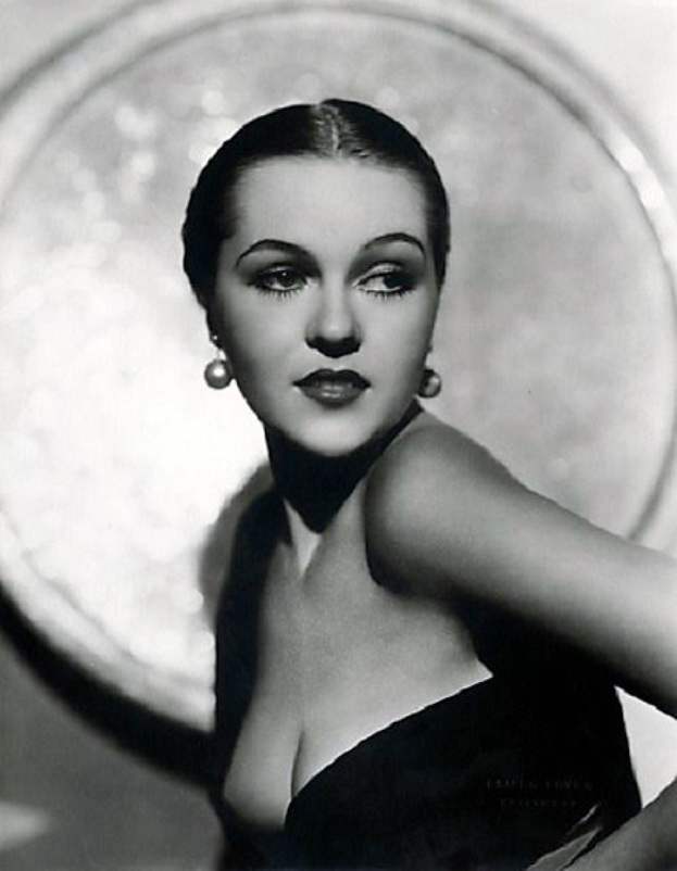 Ein Schwarz-Weiß-Foto einer Frau in einem langen Kleid im Stil der 1940er Jahre mit hohem Kragen und langen Ärmeln, gestylten Haaren, dezentem Make-up und einem selbstbewussten Ausdruck, die direkt in die Kamera blickt, mit einem unscharfen Hintergrund und Text unten.