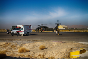 Hubschrauber auf Gras neben einem Rettungswagen mit Menschen in der Nähe, Hügel und klarer blauer Himmel im Hintergrund.