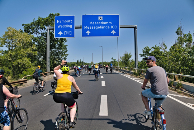 Gruppe von Radfahrern mit Helmen auf einer Straße mit einem Geländer und Bäumen auf beiden Seiten, Laternen und einem klaren blauen Himmel im Hintergrund, mit einem Schild, das eine Radtour in Hamburg anzeigt.