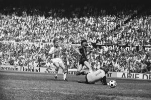 Schwarzes und weißes Foto von Männern, die Fußball auf einem Feld mit Zuschauern im Hintergrund und einem Banner "Bundesliga 1978-79 Ajax 1-1 Hannover 96" spielen.