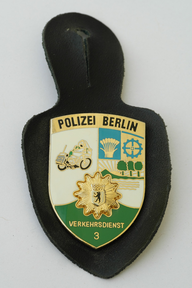 Ein schildförmiges Polizeibadgebnis mit "Polizei Berlin" in schwarzer fetter Schrift auf einem blauen Hintergrund mit einem weißen Stern und Rahmen, das auf einer Oberfläche ruht.