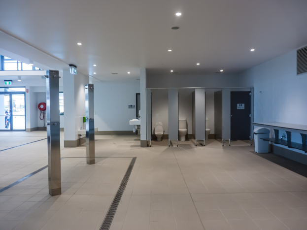 Großes öffentliches WC mit mehreren Toiletten, tragenden Säulen, einem Müllbehälter, einer Tür, einer Tafel, einem Feuerlöscher und einer Decke mit Leuchten.